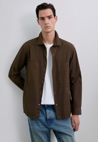 Tânăr îmbrăcat cu o cămașă deschisă, maro, cu nasturi, peste un tricou alb și blugi ușor decolorati, stând în fața unui fundal alb simplu.