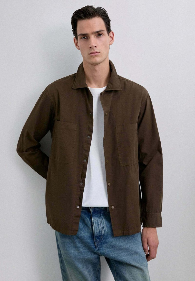 Tânăr îmbrăcat cu o cămașă deschisă, maro, cu nasturi, peste un tricou alb și blugi ușor decolorati, stând în fața unui fundal alb simplu.