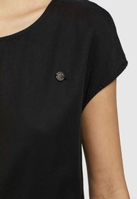 Musta puuvillapaita, jossa on pyöreä kaula-aukko ja lyhyet hihat, ja vasemmassa rinnassa on pieni pyöreä logoyksityiskohta.