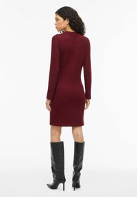 Abito aderente color bordeaux con maniche lunghe e collo alto, abbinato a stivali neri in pelle al ginocchio, con una texture liscia.