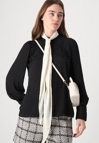 Svart bluse med blondedetaljer, knappfrigjøring og lange pufferm. Kombinert med et lyst skjerf og teksturerte rutete shorts. Liten håndveske.
