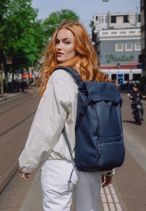 Ung kvinne med langt rødt hår iført en lys jakke og hvite bukser, bærer en stor marineblå ryggsekk på en bygate.