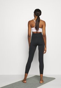 Leggings neri a vita alta con fessure laterali; abbinati a un reggiseno sportivo rosa chiaro con design incrociato sulla schiena; in piedi su un tappetino grigio.