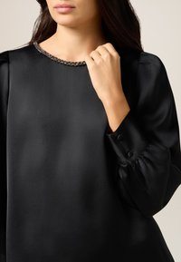 Top de satén negro con escote redondeado adornado con un borde de joyas, mangas abullonadas y puños abotonados. Textura suave, diseño elegante.