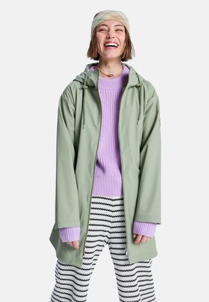 Jeune femme portant une veste de pluie verte, un pull lavande, un pantalon noir et blanc à motifs zigzag, et un foulard beige à motifs, souriant les yeux fermés.