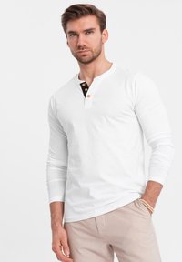 Vit långärmad henley-tröja med en svart knäppning och tre bruna knappar. Tröjan har en mjuk textur och en åtsittande design.