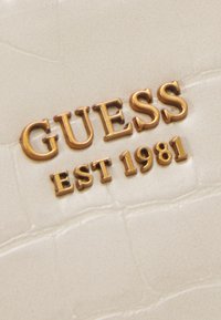 Złocone litery "GUESS EST 1981" na fakturowanym, kremowym tle ze sztucznej skóry z delikatnymi wzorami przypominającymi skórę krokodyla.