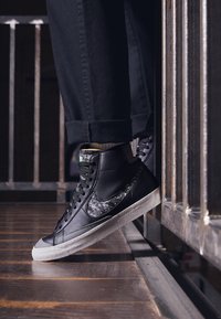 Svarta hög-top sneakers i texturerat läder med en gråmönstrad accent, vit gummisula och gröna detaljer vid kragen.