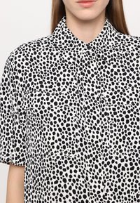Black and white short-sleeve shirt featuring a spotted pattern, with a pointed collar and two chest pockets. Soft fabric texture.