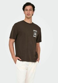 Brun bomulds T-shirt med rund hals, prydet med en hvid grafik af et kompasrose og tekst på venstre brystområde. Båret med hvide bukser.