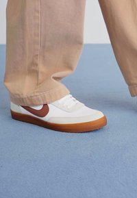 Sapatilhas brancas com detalhes castanhos e sola de borracha. Apresentam biqueira arredondada, sistema de atacadores e tecido texturizado com detalhes em couro liso.