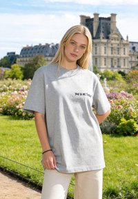 Jeune femme aux longs cheveux blonds portant un t-shirt gris avec l'inscription « YOU & ME TONIGHT », debout dans un jardin avec des bâtiments historiques derrière elle.
