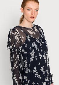 Vestido floral azul-marinho com mangas longas transparentes, tecido texturizado e padrões florais brancos. Apresenta um decote redondo e mangas ligeiramente bufantes.
