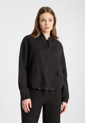 Jonge vrouw met lichtbruin haar draagt een zwarte jas met ritssluiting en bijpassende broek, staand tegen een egale lichtgrijze achtergrond.