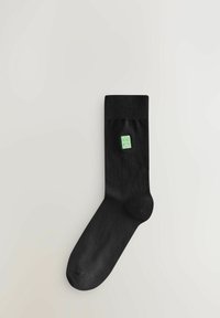 Calzino di cotone nero con polsino a coste. Presenta un piccolo logo quadrato verde vicino alla caviglia. Texture liscia, forma standard del calzino.