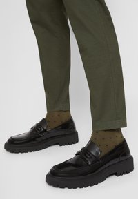 Mocassins noirs épais portés avec un pantalon vert olive et des chaussettes olive à petits pois foncés sur fond blanc.