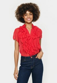 Rote Bluse mit rüschendem Frontdesign, kurzen Ärmeln und einem Kragen. Kombiniert mit dunkelblauen Jeans. Glatte Textur.