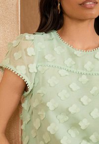 Mintgrön topp med genomskinligt tyg, prydd med texturerade blom-applikationer och en dekorativ pom-pom-detalj längs halsringningen och ärmarna.