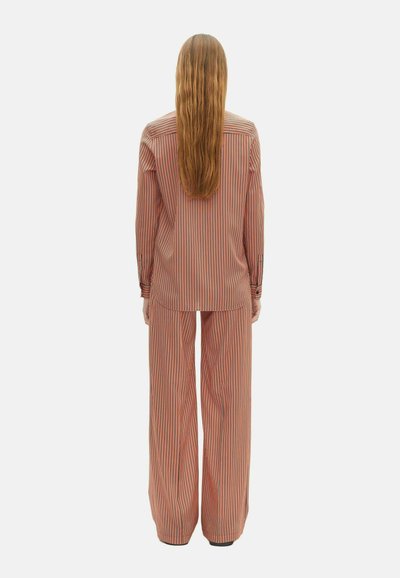 Persoon met lang, steil haar, gekleed in een losse, langmouwige shirt en brede broek met verticale oranje, zwarte en witte strepen, met de rug naar de kijker.