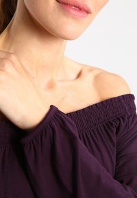 Paita off-the-shoulder dalam kain ungu tua. Memiliki leher yang berkumpul dan lengan panjang dengan manset elastis. Tekstur halus, desain santai.