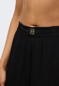 Μαύροι υφασμάτινοι παντελόνια με υφή διαθέτουν ζώνη με αγκράφα σε διπλό σχέδιο "B" με χρυσούς τόνους, που τονίζει τη γραμμή της μέσης.