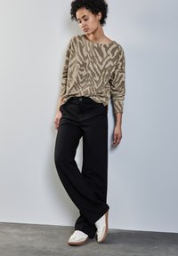 Beiger Pullover mit Zebra-Muster, schwarze weit geschnittene Hose und weiße Turnschuhe mit braunen Sohlen. Modell posiert vor einem neutralen Hintergrund.
