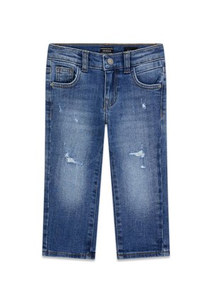 Garçon aux cheveux bouclés portant un sweat-shirt noir Guess, un jean bleu déchiré et des baskets bleu marine, debout devant un fond uni.