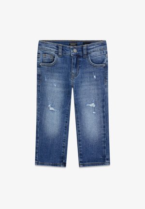 Garçon aux cheveux bouclés portant un sweat-shirt noir Guess, un jean bleu déchiré et des baskets bleu marine, debout devant un fond uni.