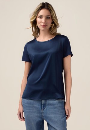 Haut bleu marine à manches courtes, en satin lisse, avec un col arrondi et des fentes sur les côtés, associé à un jean en denim bleu clair.