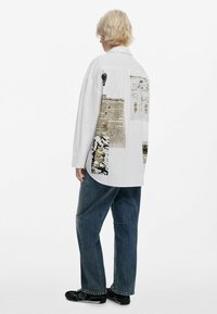 Chemise blanche surdimensionnée avec des motifs graphiques, incluant du texte et des illustrations, au dos. Portée avec un jean bleu ample et des chaussures noires.