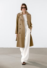 Brązowy płaszcz trenczowy z podwójnym zapięciem i naramiennikami, noszony na białej bluzce i szerokich spodniach. Osoba ma również na sobie ciemne okulary przeciwsłoneczne i sandały.