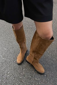 Tan suede kniehoge laarzen met een licht gebogen schacht, een rits aan de zijkant en een platte rubberen zool, geplaatst op een gestructureerd asfaltoppervlak.