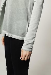 Maglia grigia chiaro a maniche lunghe realizzata in un morbido tessuto testurizzato. Design a taglio con una fessura laterale e un logo sull'orlo. Indossata con pantaloni neri.