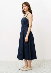 Marineblaues ärmelloses Kleid mit figurbetontem Oberteil und fließendem Rock, hergestellt aus leichtem Stoff; kombiniert mit hellen Schnürschuhen.