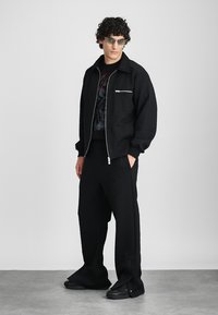Black textured jacket with a silver zip pocket, paired with matching black pants featuring side snaps. Worn with black sneakers and tinted sunglasses.