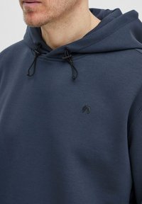 Jūras zila kapuce ar auklu, kurai ir mazs melns logotips uz krūtīm. Gluda tekstūra ar brīvu piegriezumu.