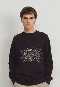 Sudadera negra con un diseño circular de tachuelas que presenta la palabra "HUGO" en el centro. Tejido suave y corte relajado.
