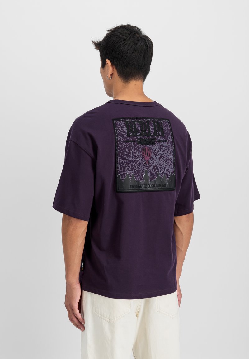 T-shirt en coton violet avec un grand imprimé graphique au dos, présentant un design de carte et une silhouette de groupe. Manches courtes, coupe décontractée.