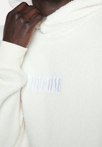 Weißer Fleece-Hoodie mit weicher Textur und einem erhabenen blauen "VOLCOM"-Logo. Die Bündchen sind gerippt, und die Kapuze ist gemütlich gestaltet.