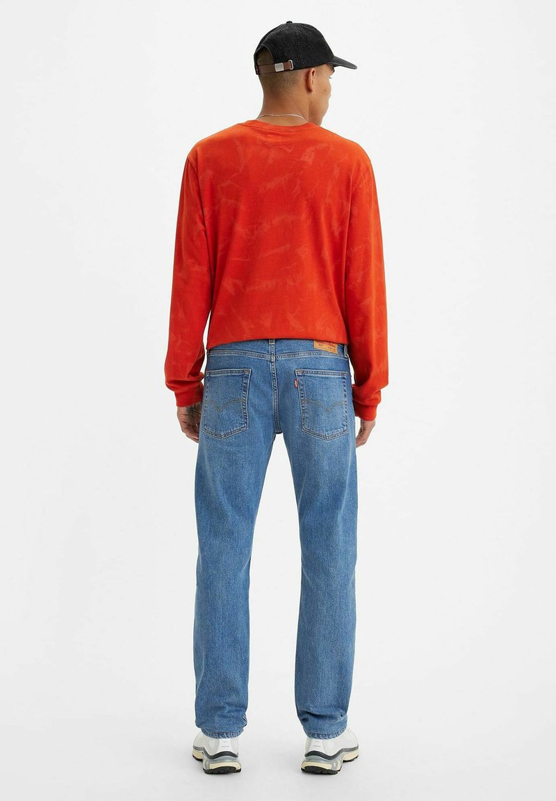 Czerwona koszula z długim rękawem o teksturowanym wzorze, w połączeniu z jasnoniebieskimi dżinsami. Czarna czapka i sneakersy z białą podeszwą dopełniają wygląd.