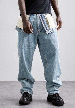 Mężczyzna w jasnobłękitnych luźnych dżinsach z dużymi naszywanymi kieszeniami i czarnych masywnych skórzanych butach, stojący na szarej podłodze.