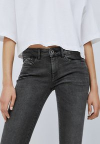 Svarta denimjeans med en figurnära design, med blyknappar, femfickorsstil och subtilt blekt mönster. En croppad vit topp är delvis synlig.