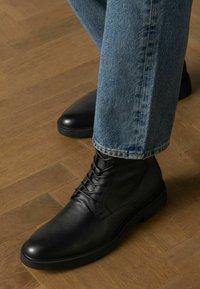 Bottines en cuir noir avec un bout arrondi et un design à lacets, assorties à un jean en denim bleu clair, sur un sol en bois.