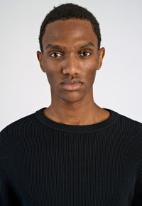 Schwarzer gerippter Pullover mit rundem Halsausschnitt, aus einem strukturierten Stoff und mit einer taillierten Silhouette, getragen von einem Modell mit kurzen Haaren.