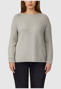 Vrouw in een lichtgrijze gebreide trui met lange mouwen en ronde halslijn, gecombineerd met zwarte broeken, die tegen een effen achtergrond staat.