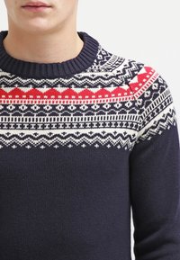 Man som bär en marinblå stickad tröja med vitt och rött nordiskt mönster över bröstet och axlarna, ribbad rund halsringning.