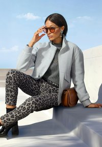 Frau trägt eine graue Bomberjacke, ein dunkelgraues Oberteil, Leopardenmuster-Hosen, schwarze Stiefel, Sonnenbrille und auffällige Ohrringe, sitzt im Freien auf Treppen.