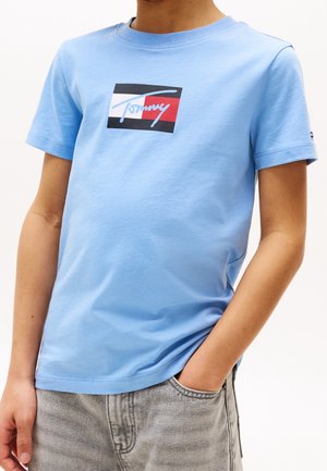 Barn iført en lyseblå kortærmet t-shirt med et rødt, hvidt og sort logo og grå cowboybukser med den ene hånd i lommen.