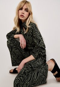 Green and black patterned outfit featuring a leaf design, long sleeves, and loose fit. Worn with dark sandals. Smooth fabric texture.