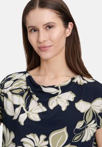 Bluse mit floralem Muster in Marineblau, mit einem drapierten Ausschnitt, kurzen Ärmeln und großen creme- und salbeigrünen Blumen. Glattes, leichtes Material.